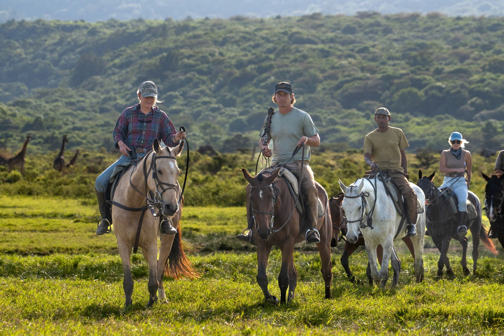 Kilimanjaro Elephant Ride, Arusha National Park, Tanzania – elefant i högt gräs tittar mot kameran, medan fem ryttare till häst på ridsafari i bakgrunden betraktar elefanten i ett grönt och frodigt landskap.