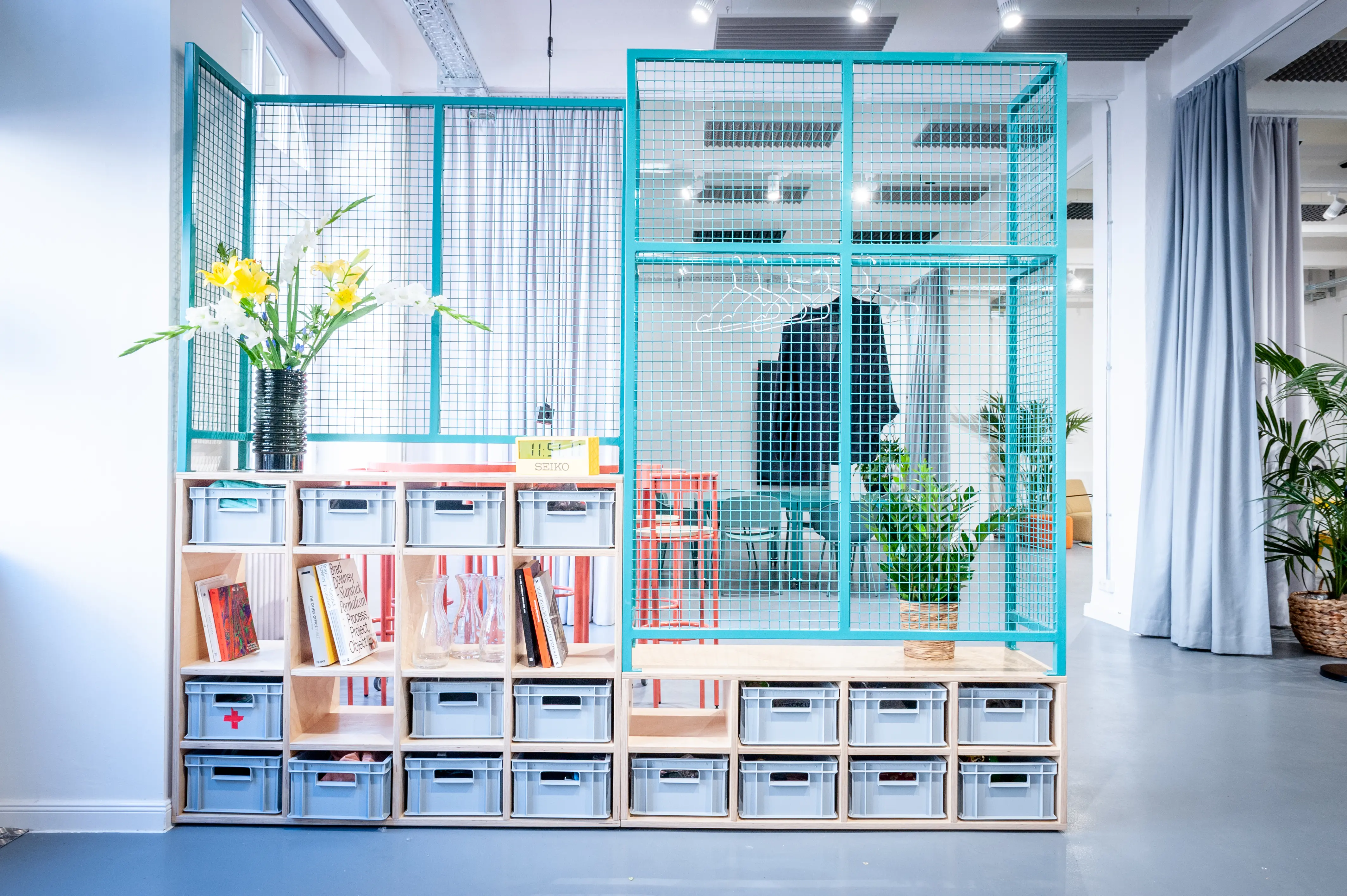 Room-divider module at HEY CONTENT STUDIO combining a turquoise wire mesh frame with a plywood shelf base holding grey storage crates, books and flowers.