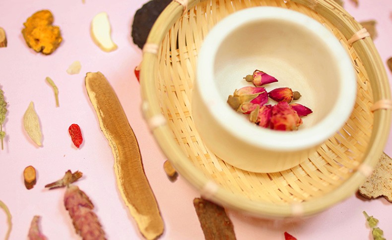 A small white bowl containing a few dried flower petals sits inside a light-colored woven bamboo basket. The basket rests on a light pink surface. Surrounding the basket are various dried plant parts, including thin, curled stems, flat, disc-shaped pieces, and small, round seeds. The dried plant materials are scattered across the surface, with some pieces lying near the basket and others further away. The background is a uniform light pink color.