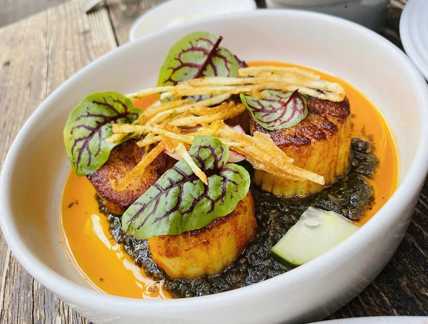 White plate with roasted carrots served on green sauce, topped with almonds and fresh herbs, held by a hand over a dark table.