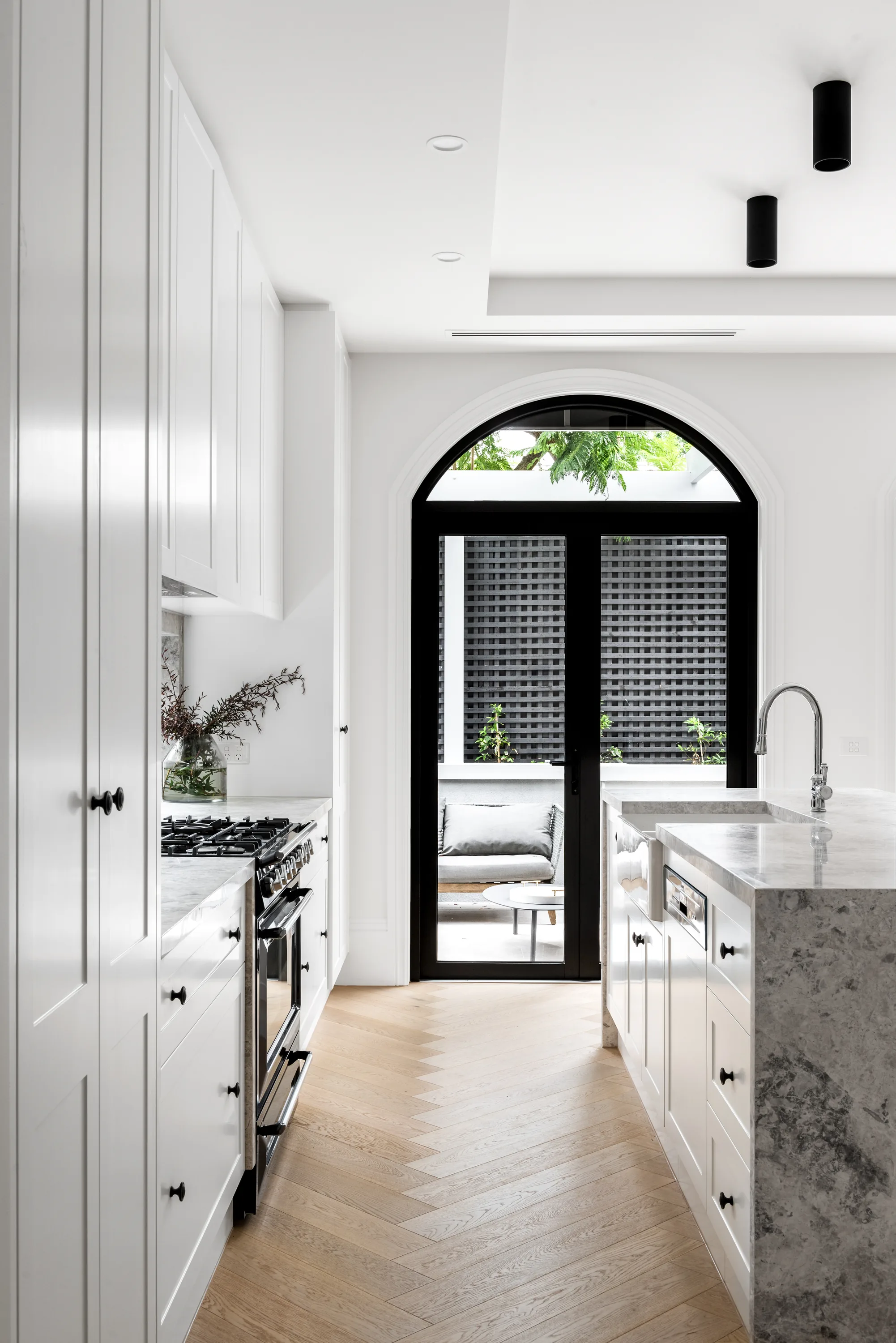Galley kitchen with white shaker cabinetry, a freestanding cooker, grey-marble island with chrome tap and herringbone floors, leading to an arched black-framed door to the courtyard