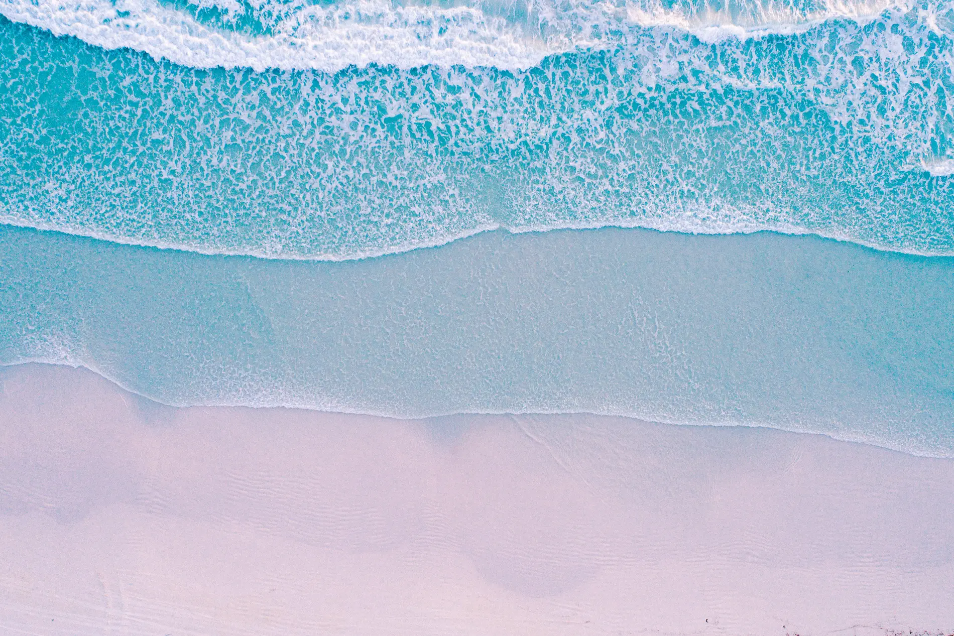 Aerial looking straight down on turquoise surf washing onto pale pink sand