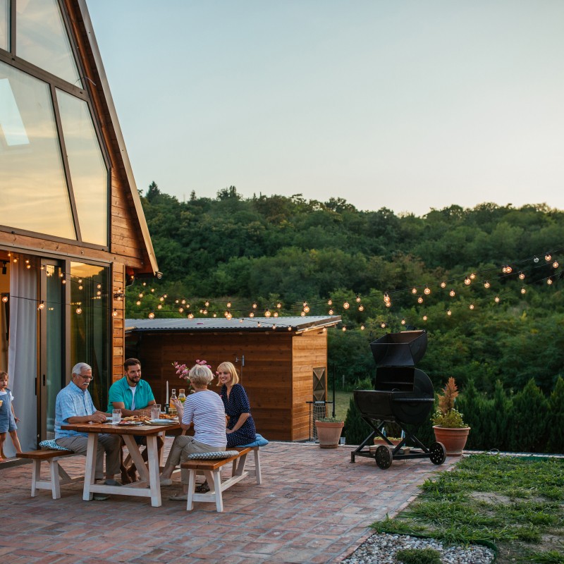 Terrasse de chalet avec famille dînant, guirlandes lumineuses et vue sur forêt au crépuscule.