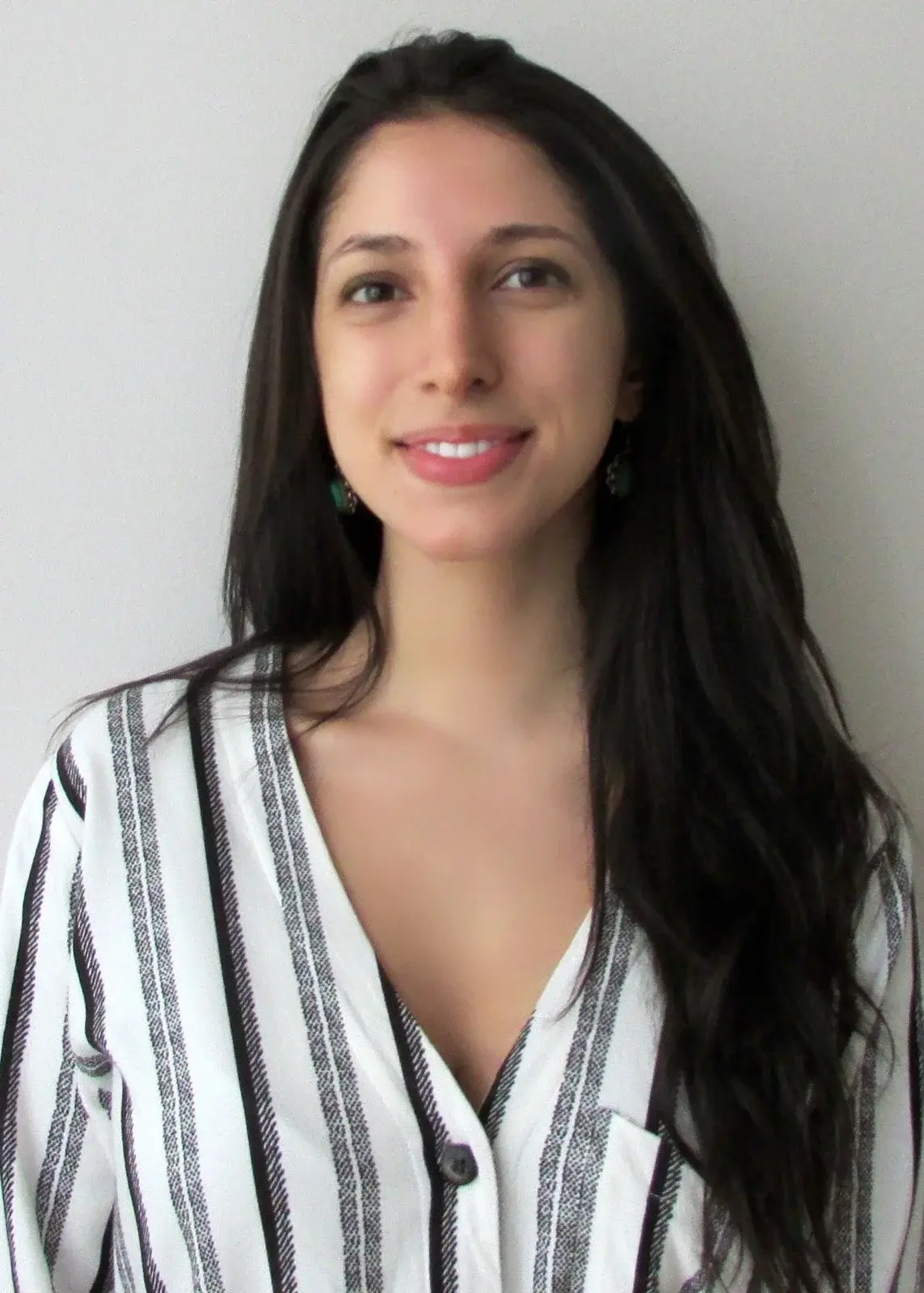 A woman with long dark hair smiles warmly at the camera, wearing a white and black vertically striped shirt against a plain light-colored background.