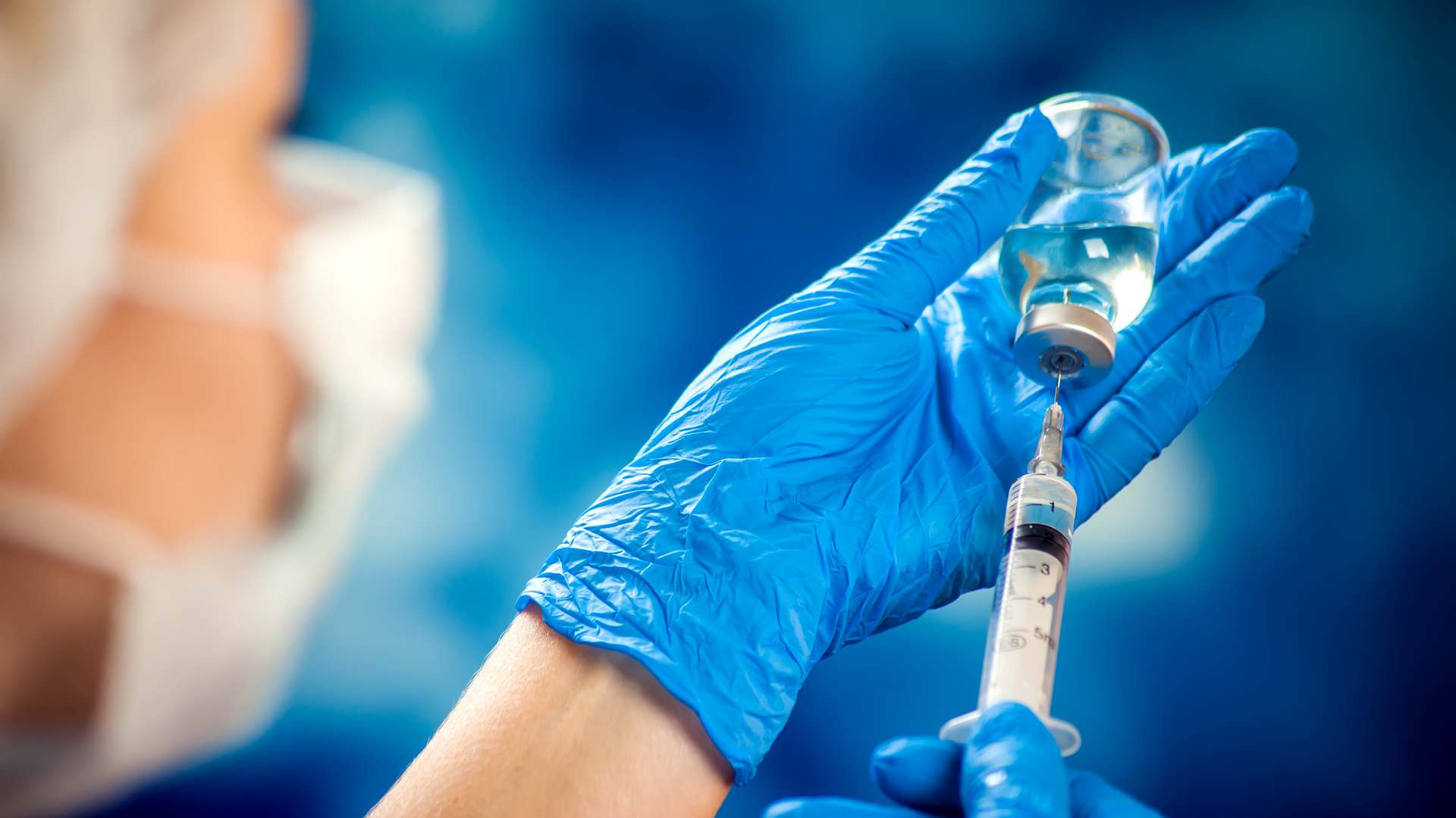 Blue-gloved hands drawing liquid from a glass vial into a syringe, with a masked clinician blurred behind