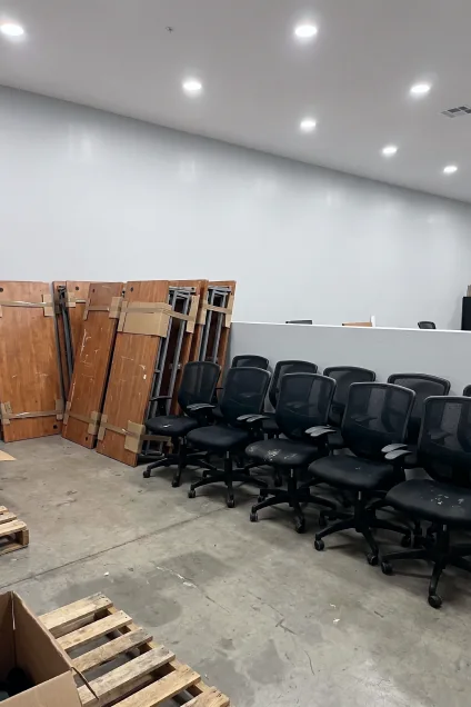 Una fila de sillas de oficina con respaldo de malla negra alineadas junto a tableros de escritorios de madera desmontados y apoyados contra la pared.