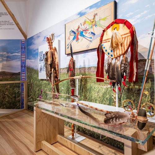 Museum display featuring Native American artifacts, including feathered headdresses, a bow, and traditional artwork on a backdrop of grasslands.