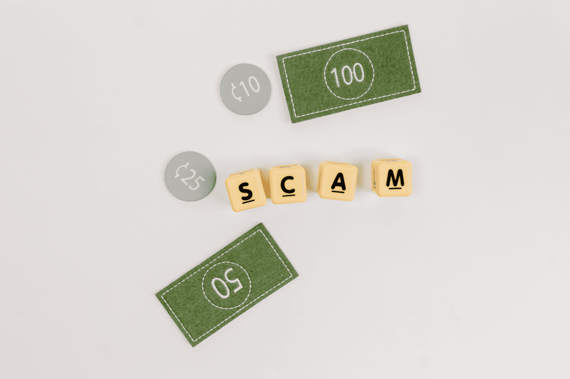 An arrangement of yellow lettered blocks spelling 'SCAM' placed on a white background alongside green felt currency notes labeled 100 and 50, and gray circular tokens labeled 10 and 25, symbolizing financial deception.