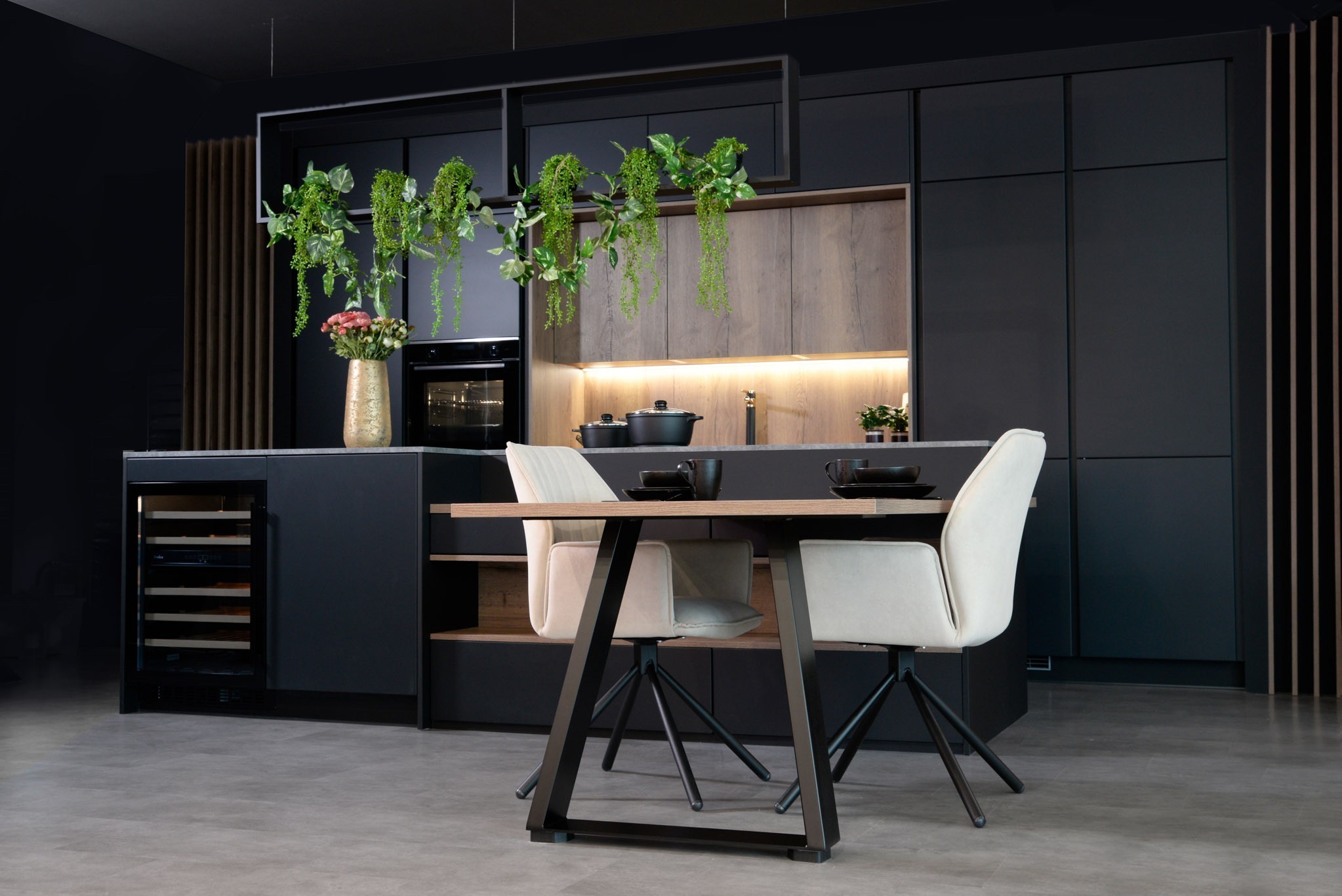 Elegante DAN Küchenzeile in dunklen Tönen mit integriertem Esstisch und bequemen Stühlen, ergänzt durch hängende Pflanzen.