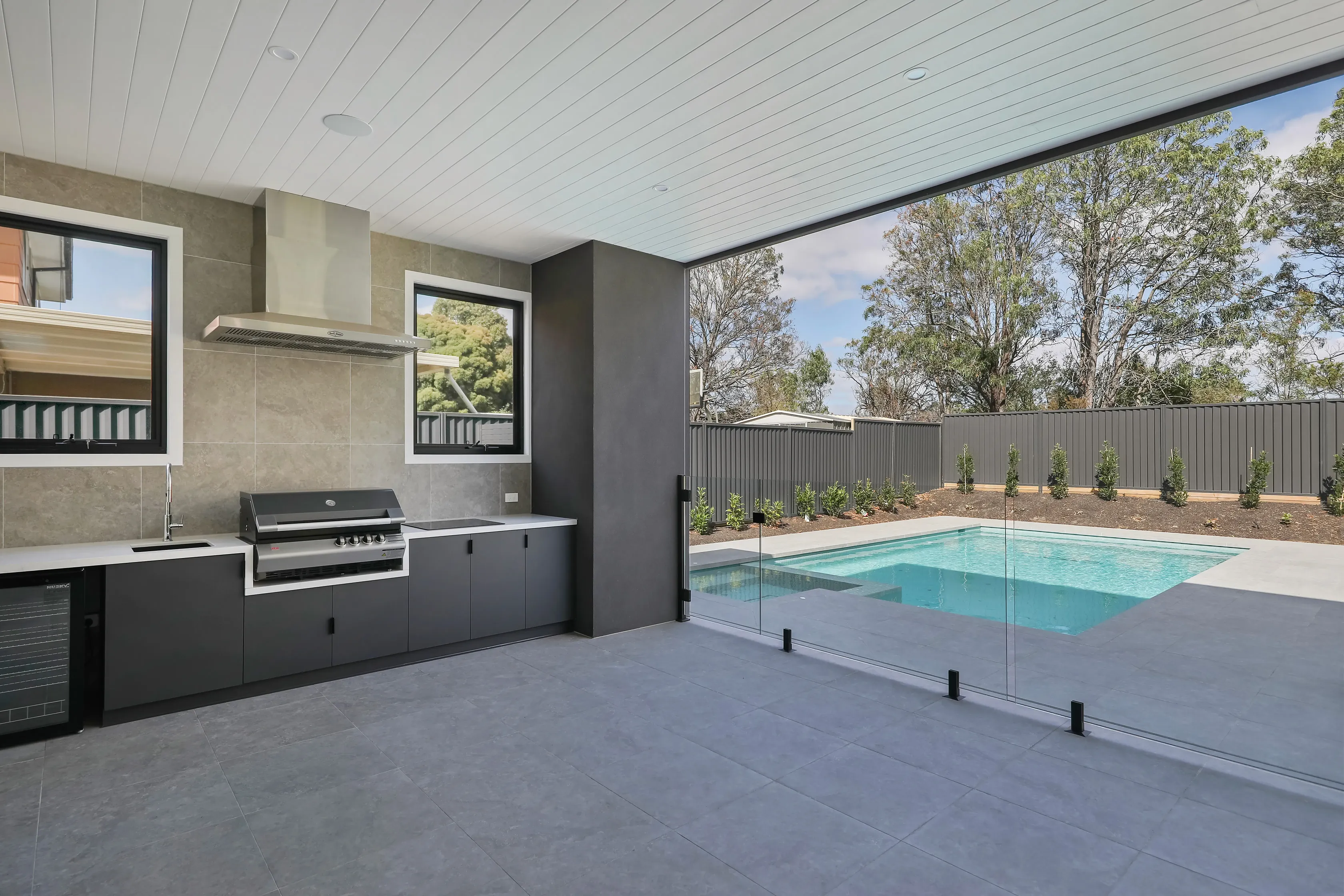 Covered alfresco area at Mount Waverley Residence with an outdoor kitchen and barbecue overlooking a glass-fenced pool and established gums