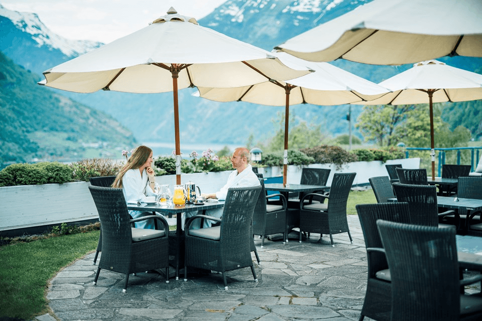 A picturesque outdoor dining area with wicker chairs and tables under large white umbrellas, set against a stunning backdrop of scenic mountains and lush greenery.