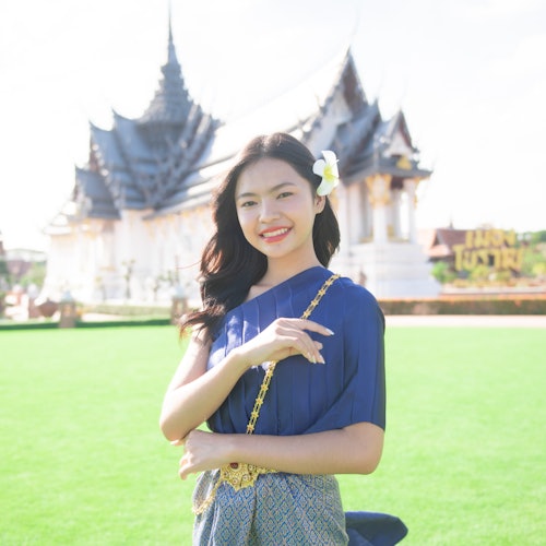 一位身着传统服饰的女子站在绿草地上，微笑着，头上戴着一朵花，背景是一座华丽的寺庙。