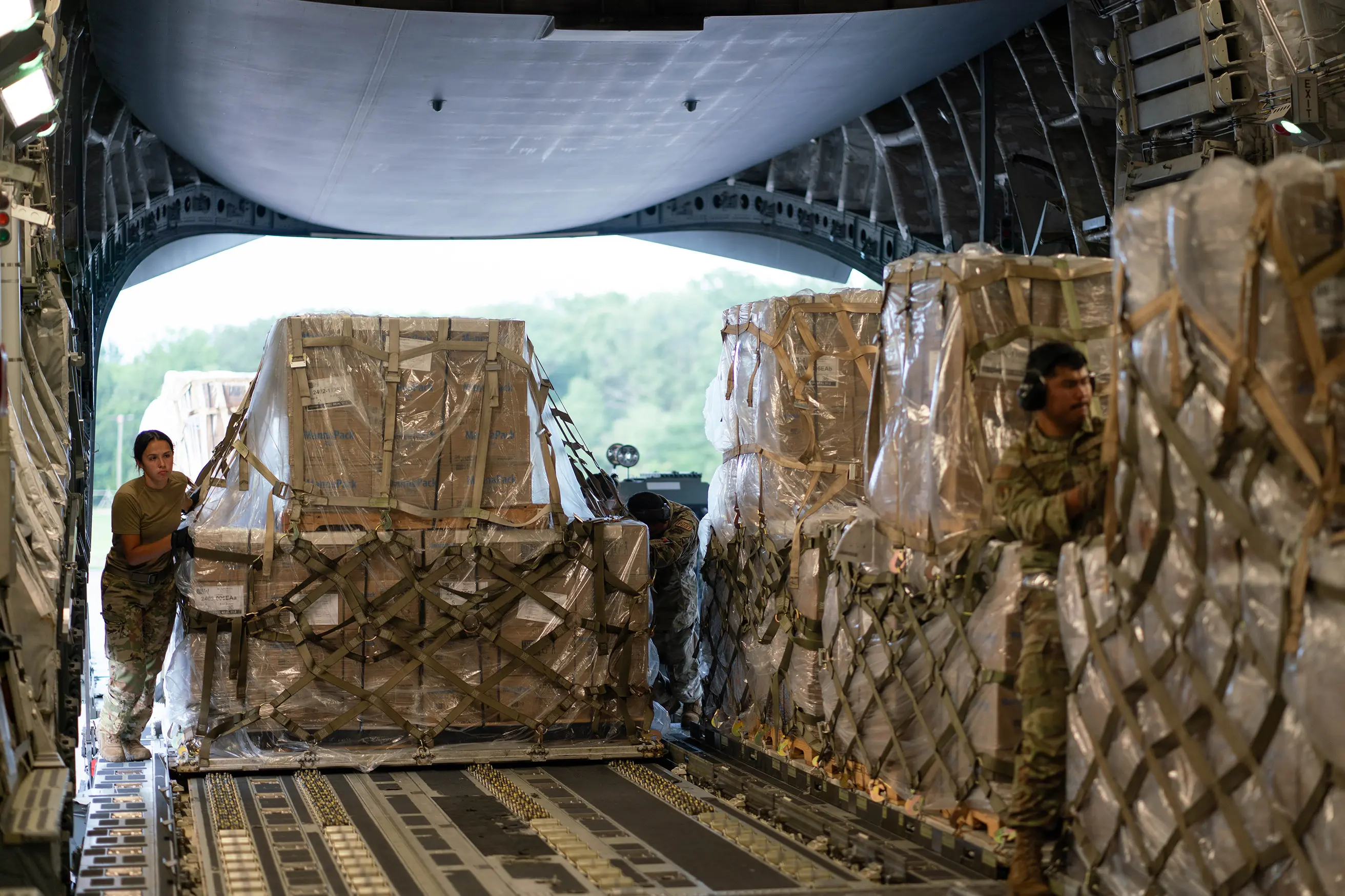 Military personnel securing large cargo pallets inside an aircraft, with straps and packaging in a spacious interior.