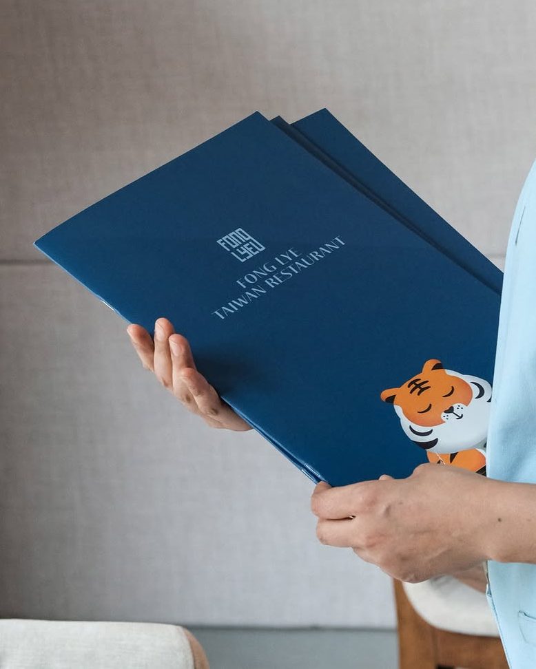 A person holding the menu of Fong Lye Taiwanese Restaurant, featuring a blue cover with a cartoon tiger and the restaurant's name prominently displayed.
