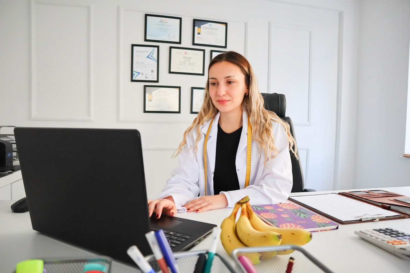 A registered dietitian in a white coat working at a laptop for a virtual nutrition consultation, with fresh bananas on the desk