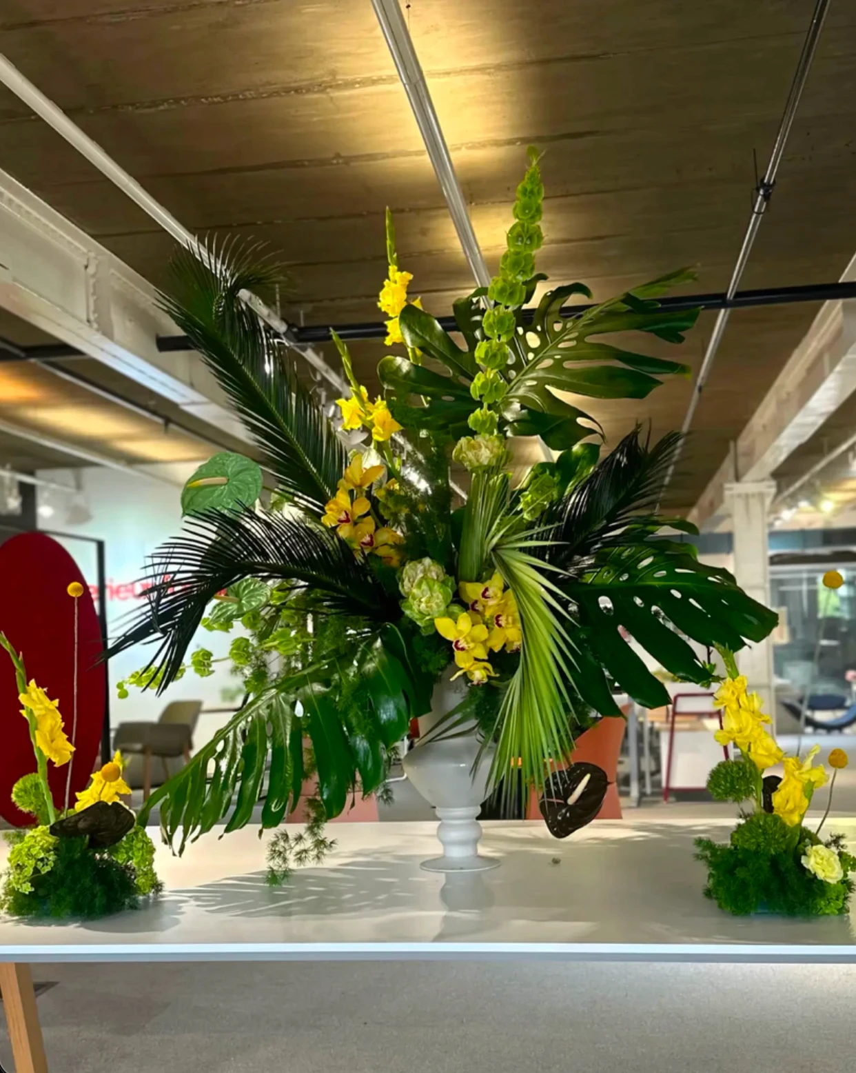 Arreglo floral tropical con hojas de monstera, orquídeas amarillas y palmas en jarrón blanco sobre mesa moderna.