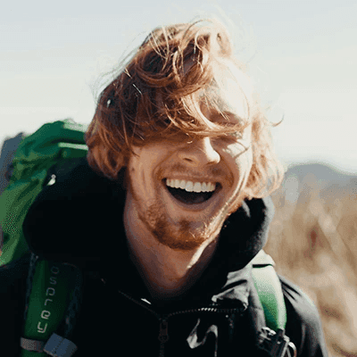 Smiling man with windswept red hair outdoors, wearing a black jacket and green backpack. Bright sunlight and a blurred natural background convey joy and adventure.