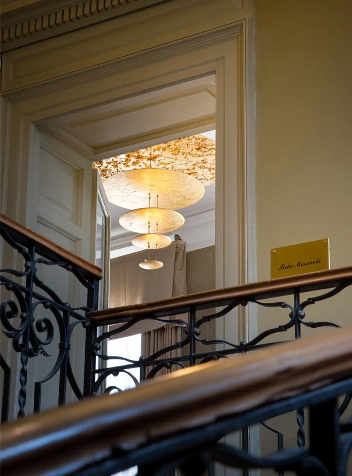 Macchina della Luce de Catellani & Smith en entrada al subir escalera con pasamanos de madera y hierro fundido negro. Iluminación de lujo | ábc concept