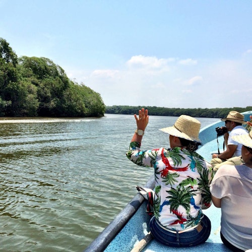 Những người đội mũ đi thuyền qua sông, vẫy tay chào bờ sông xanh mát đầy rừng dưới bầu trời trong xanh, nắng đẹp.