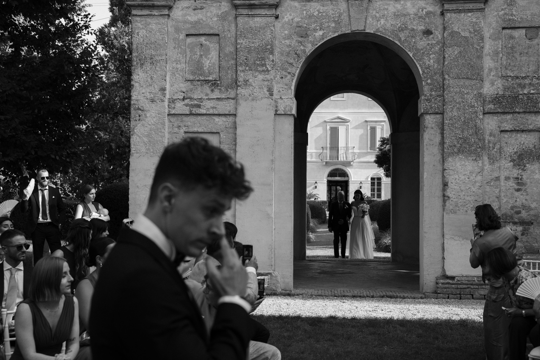 Sposi attraversano l'arco di Villa Negri, reportage matrimoniale in bianco e nero con architettura e invitati sullo sfondo