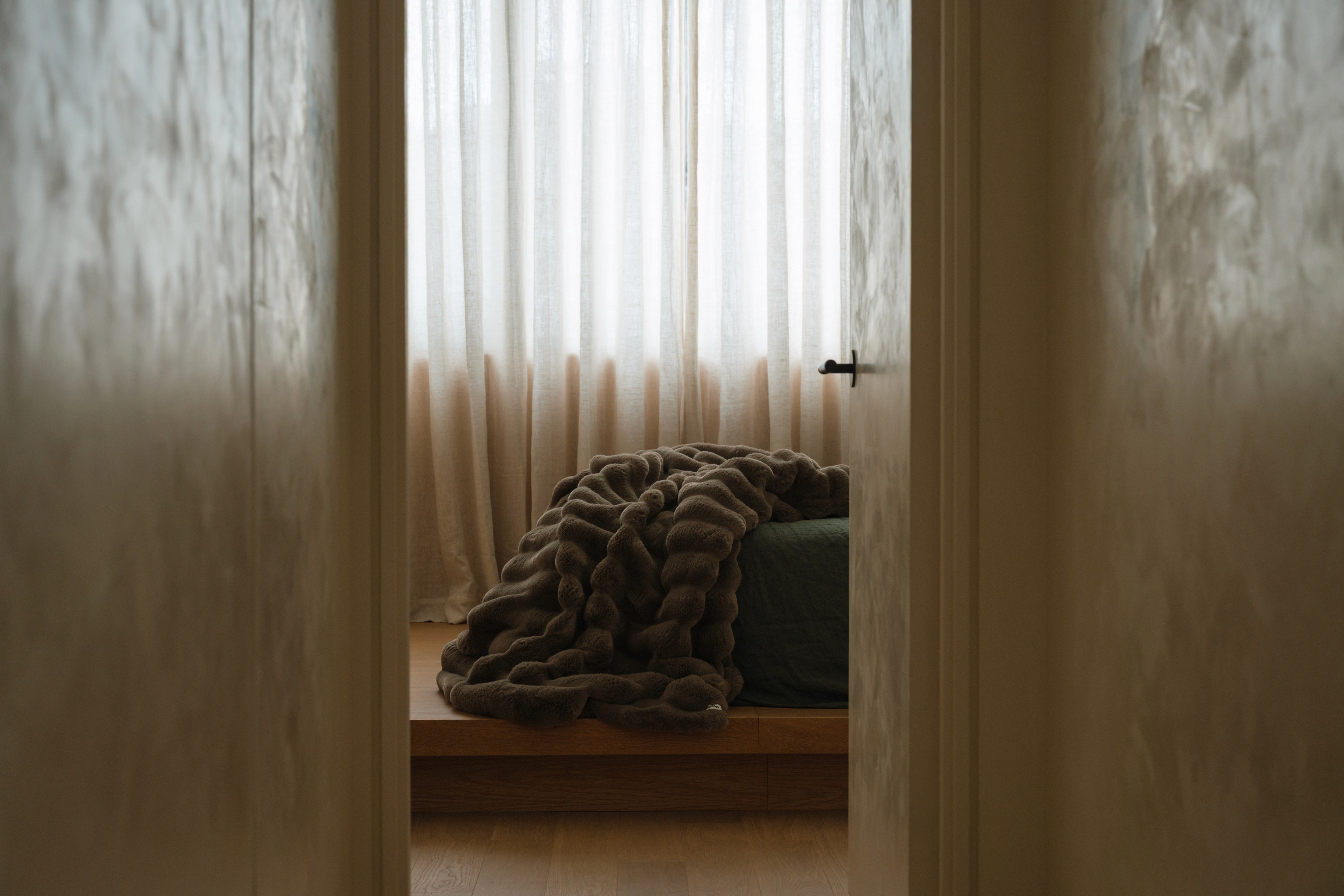 A soft cream-colored Normy blanket with a visible brand label is draped inside a decorative bathroom sink. The scene also includes bath products around the sink and a mirror reflecting the cozy setup, creating a playful and inviting atmosphere.
