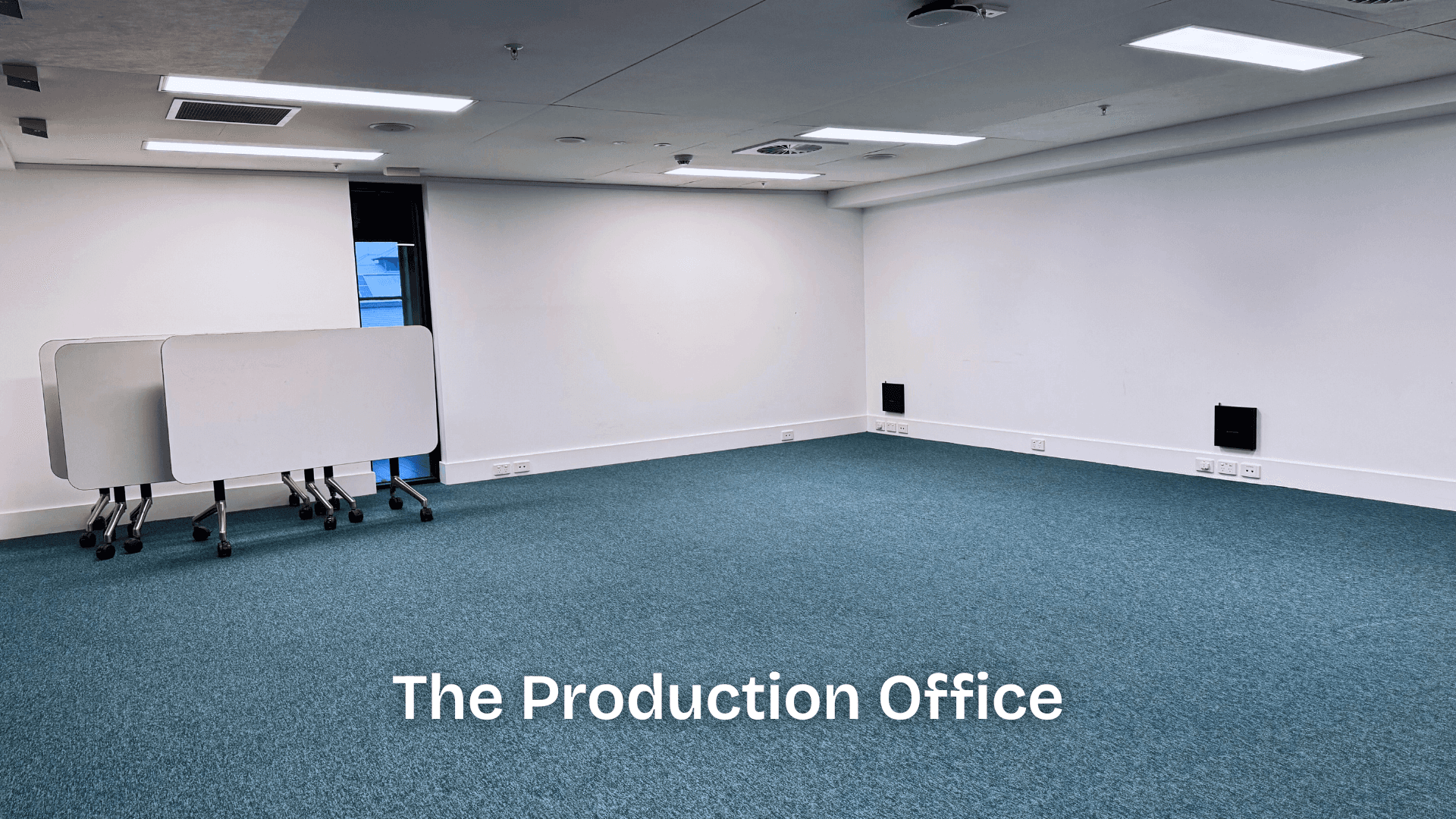 A large, empty room with blue carpet, white walls, and stackable foldable tables, arranged as a flexible production workspace.
