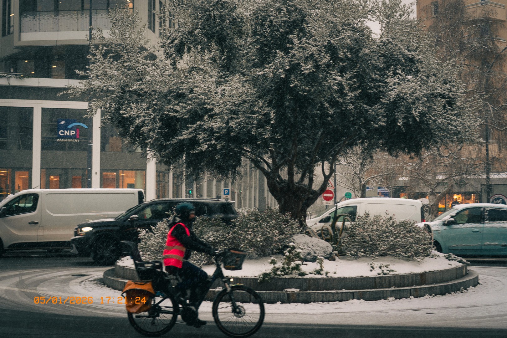 Un cycliste en gilet haute visibilité traverse le rond point Victor Hugo, avec des bâtiments modernes, des véhicules en arrière-plan et un arbre enneigé au centre de la scène.