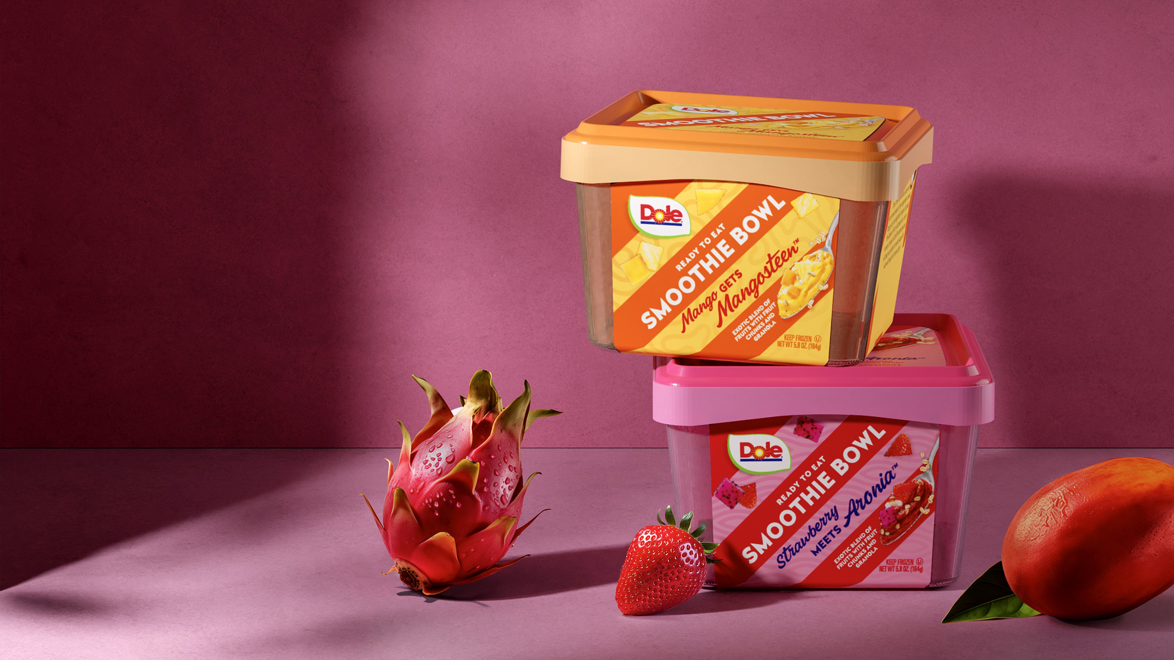 Two Dole Smoothie Bowl containers, one Mango variety and one Strawberry, are stacked on a pink surface with a dragon fruit, strawberry, and mango placed nearby.