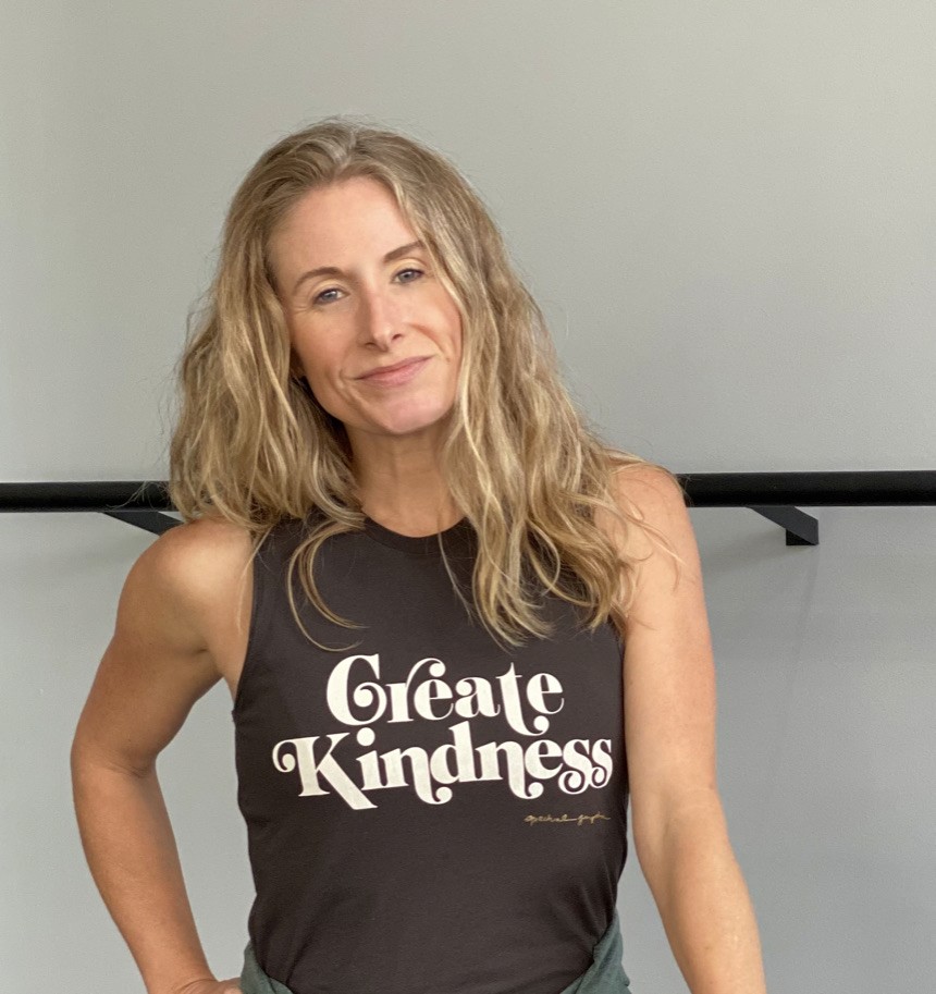 A person with long hair smiles while wearing a black sleeveless shirt that says "Create Kindness" in white text, standing against a gray background.