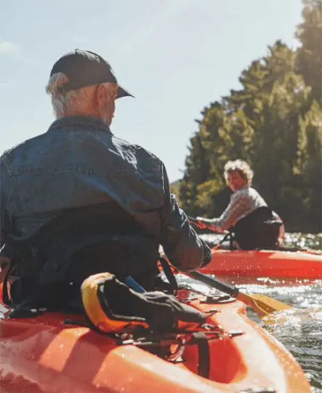 Two people kayaking on a sunny day, with trees in the background and one person in the foreground facing away.