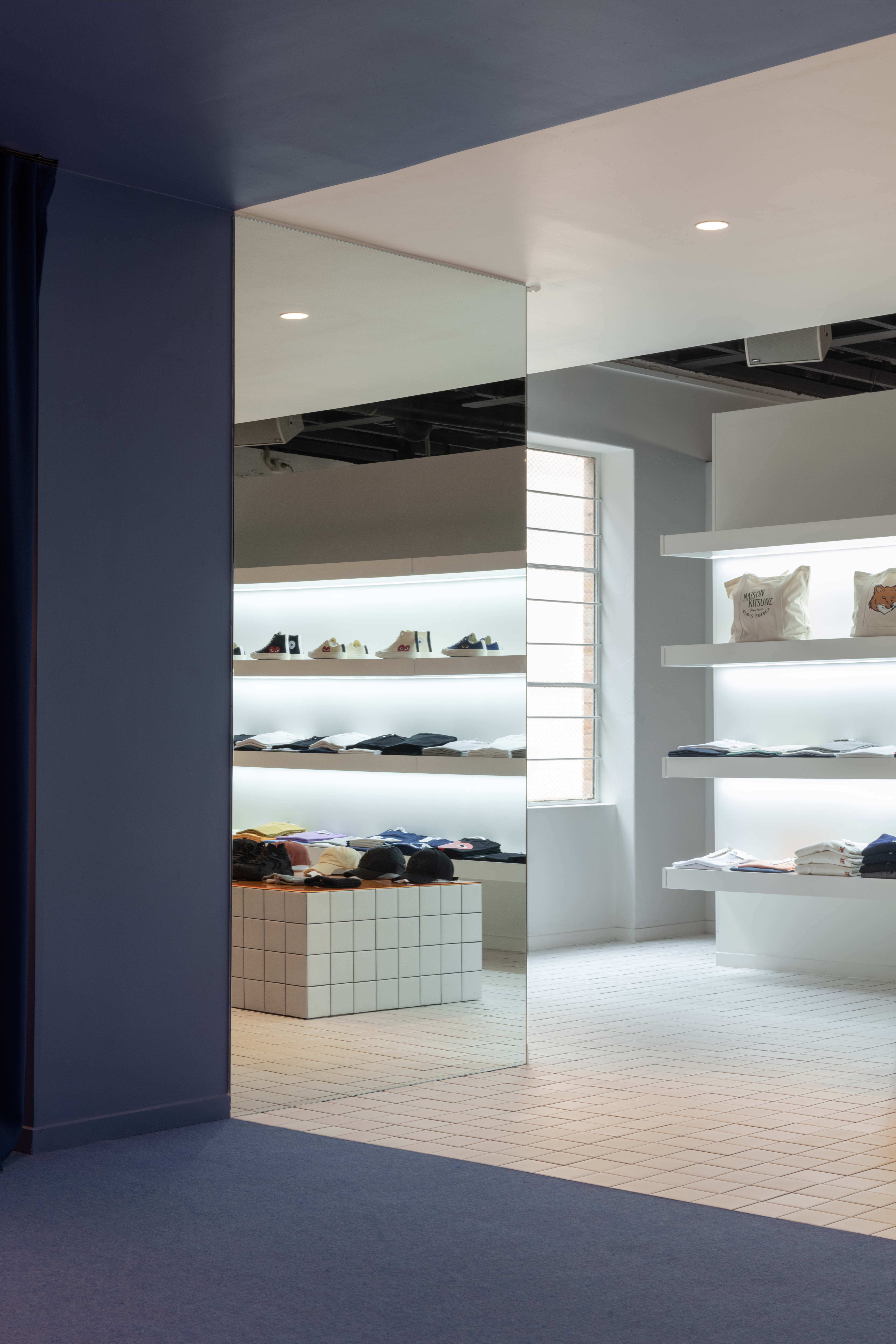 Interior transition within the 199A® space, where deep blue walls and curtains give way to a bright tiled retail zone with floating shelves, mirrored surfaces, and softly lit product displays.