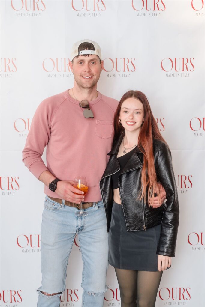 A couple posing in front of a white step-and-repeat; the man in a mauve pullover and the woman in a black leather moto jacket, capturing the relaxed yet refined dress code of a mountain event.