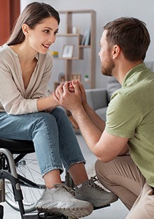 A man kneeling and holding the hands of a smiling woman sitting in a wheelchair.