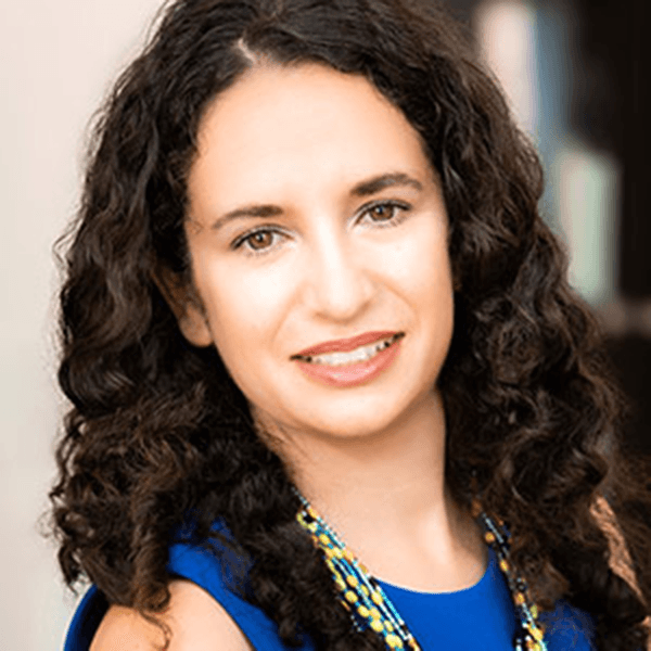 A woman with curly hair smiles warmly, wearing a blue top and a colorful necklace, set against a blurred background.