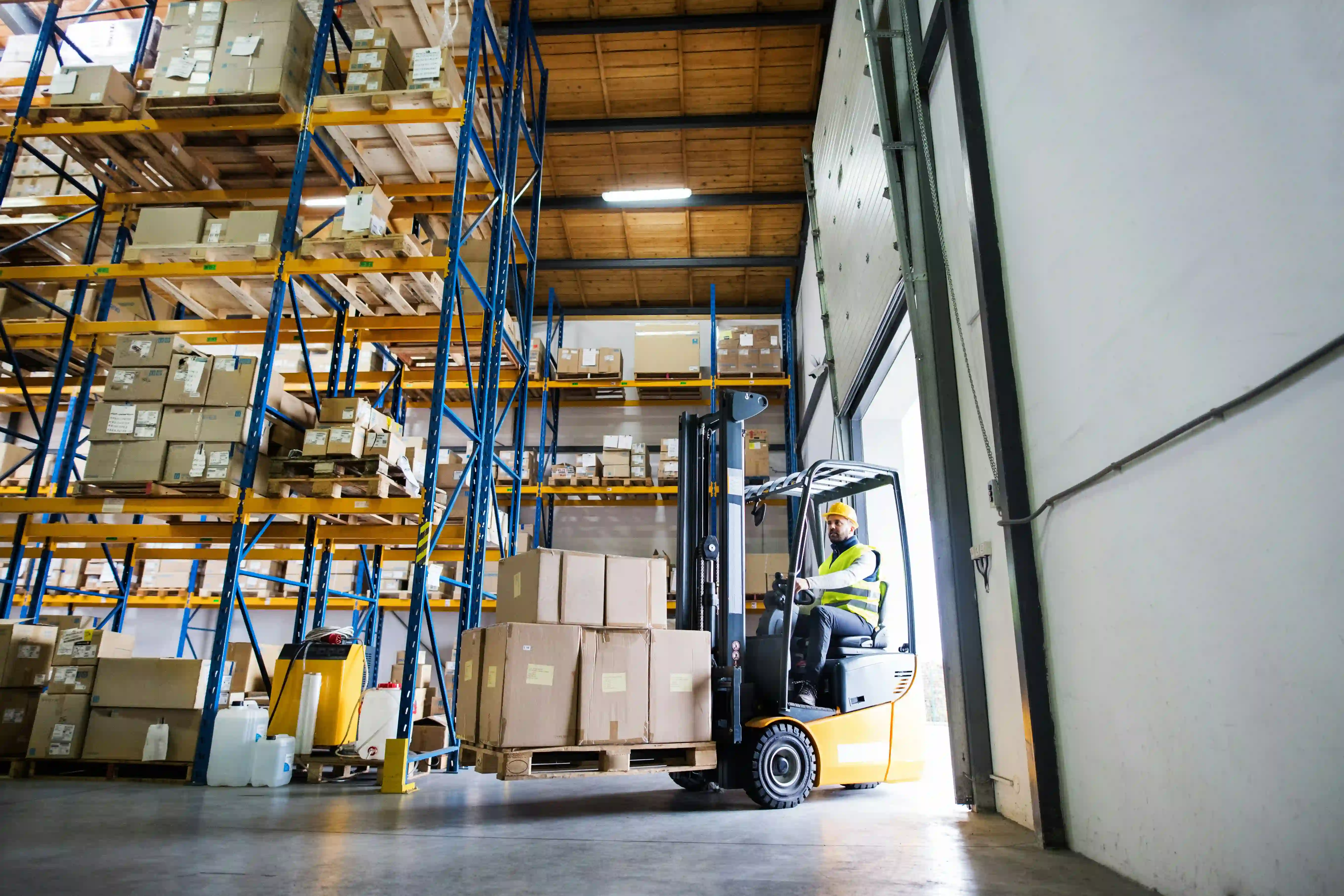 a man in a yellow forklift carries a pallet full of boxes into a warehouse via an open door