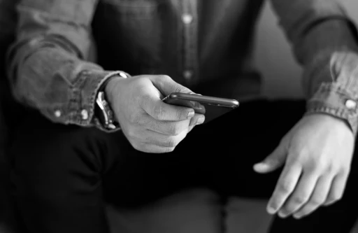 Man sitting with smartphone in hand, wearing a denim shirt and wristwatch, close-up of hands using mobile device