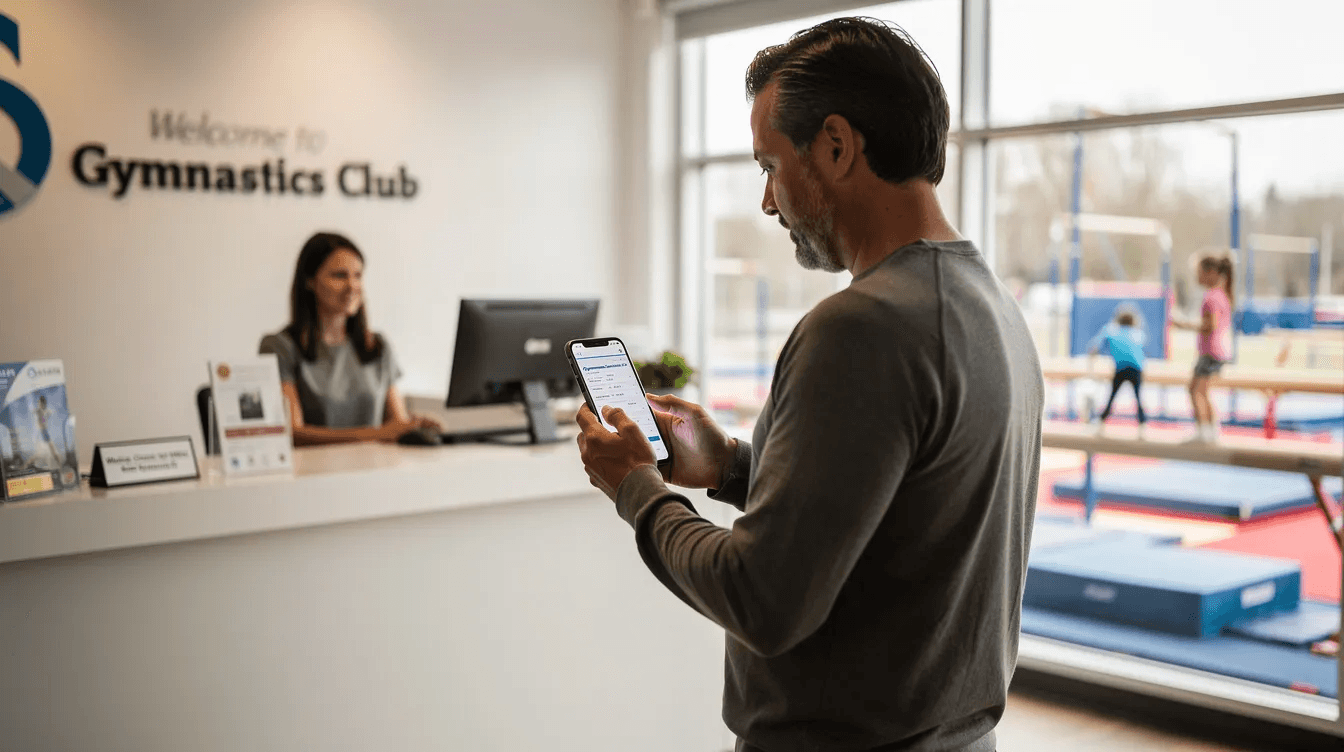 A parent is sitting in a gymnastics club reception area, using a smartphone to check the class schedule, which is part of the club management software. The environment is lively, reflecting the excitement of gymnastics training with posters of gymnasts and equipment like balance beams and uneven parallel bars visible in the background.