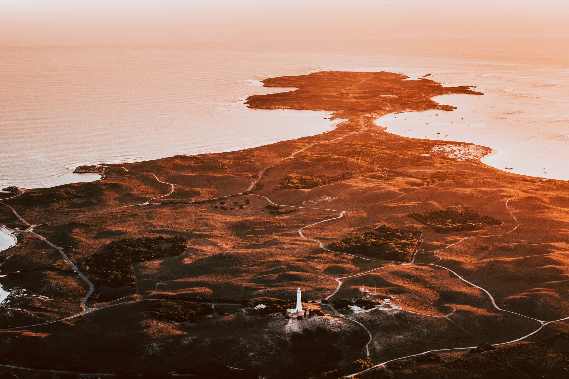 Aerial at sunset over a low island with a lighthouse and pale surrounding water
