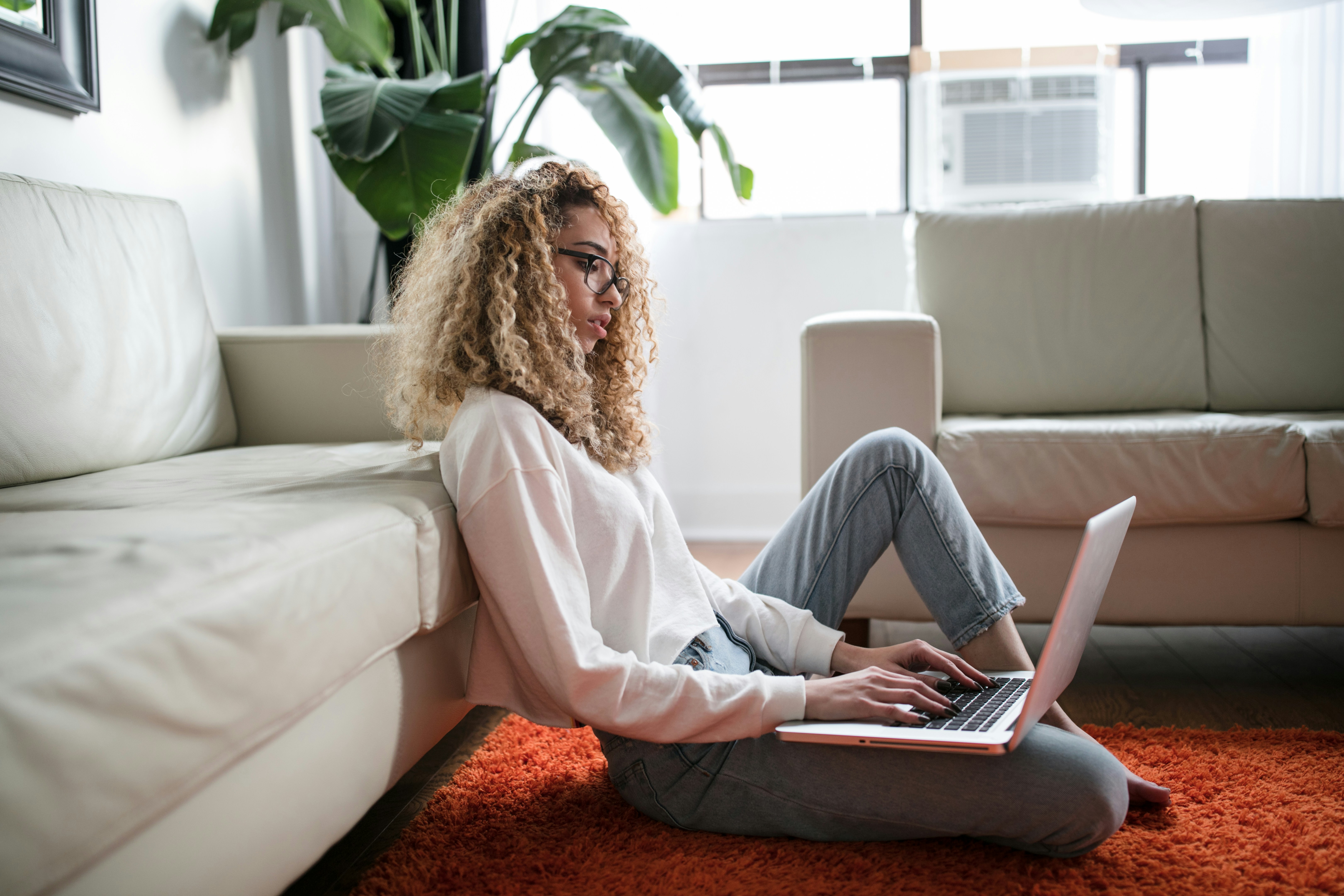 A woman sitting on the floor looks down at their open laptop.