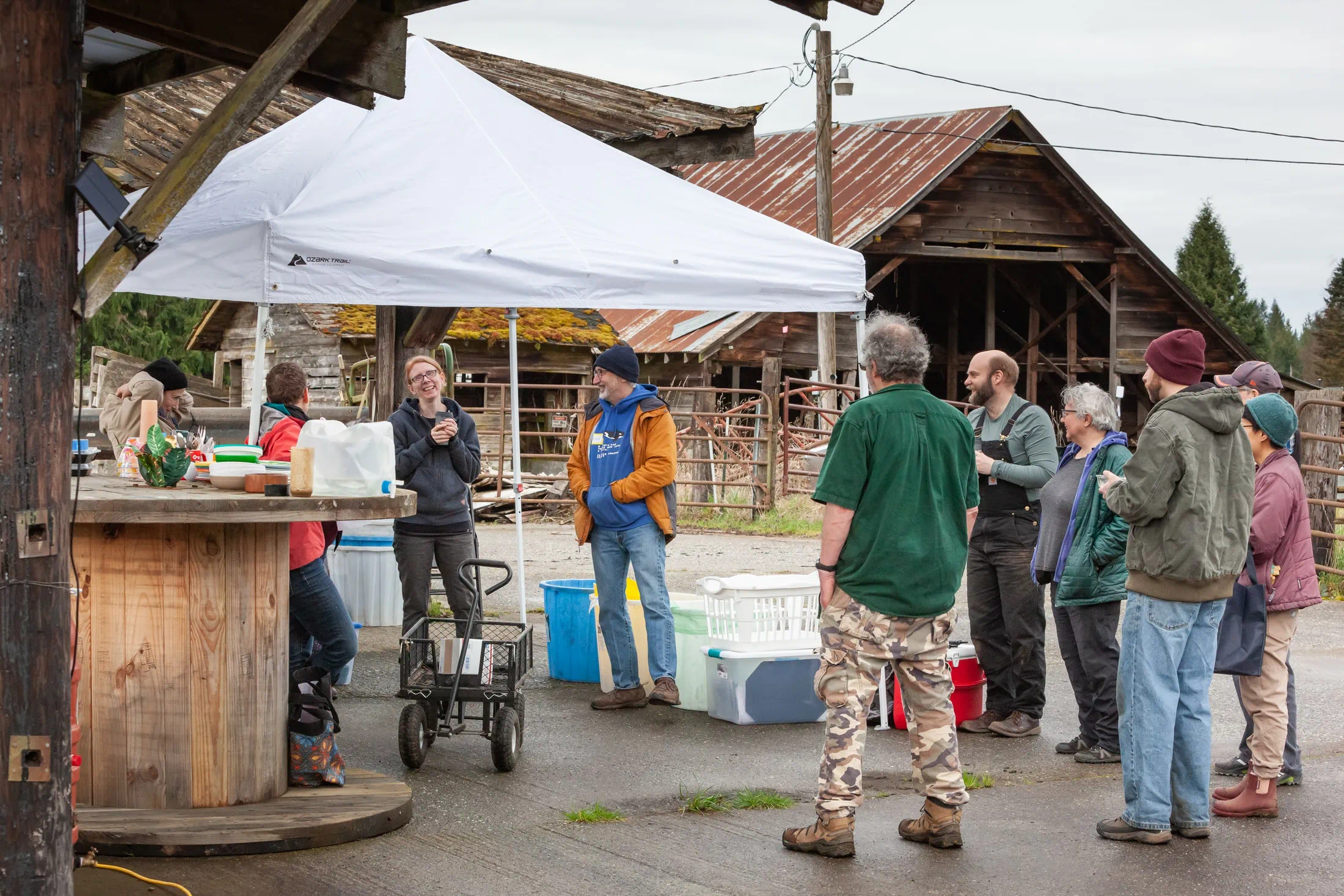 Small group of people gathered outdoors near a rustic barn and canopy tent during a community work day.