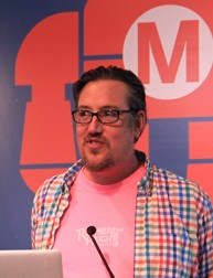 A photo of a person giving a lecture while sitting in front of a panel on which a red robot with an M is drawn.