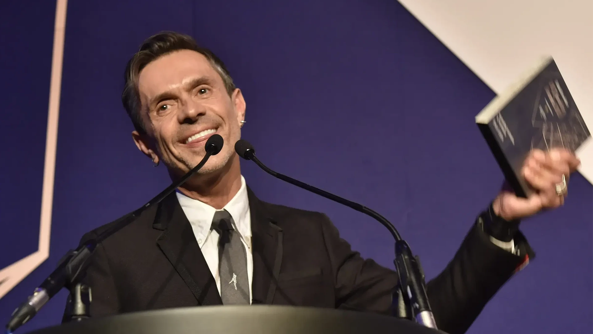 Shaun Leane smiling at a podium, holding up his Outstanding Contribution to the Industry award trophy.