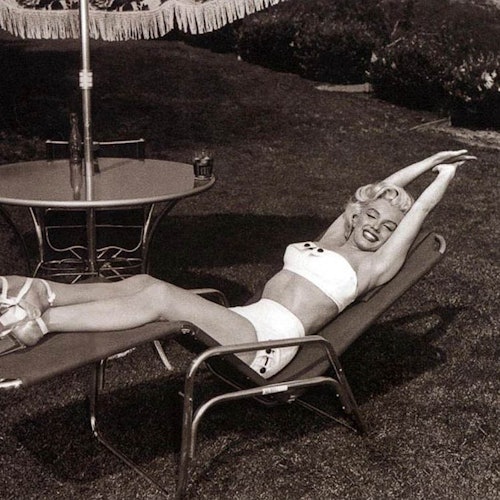 一位女士穿着两件式泳装,躺在带流苏伞的圆桌旁边的沙滩椅上,双臂伸展在头顶上微笑。