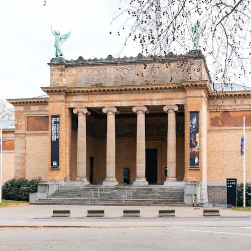 这是一座拥有新古典主义外观的历史建筑，设有六根柱子，屋顶上有雕像，入口两侧悬挂着横幅。