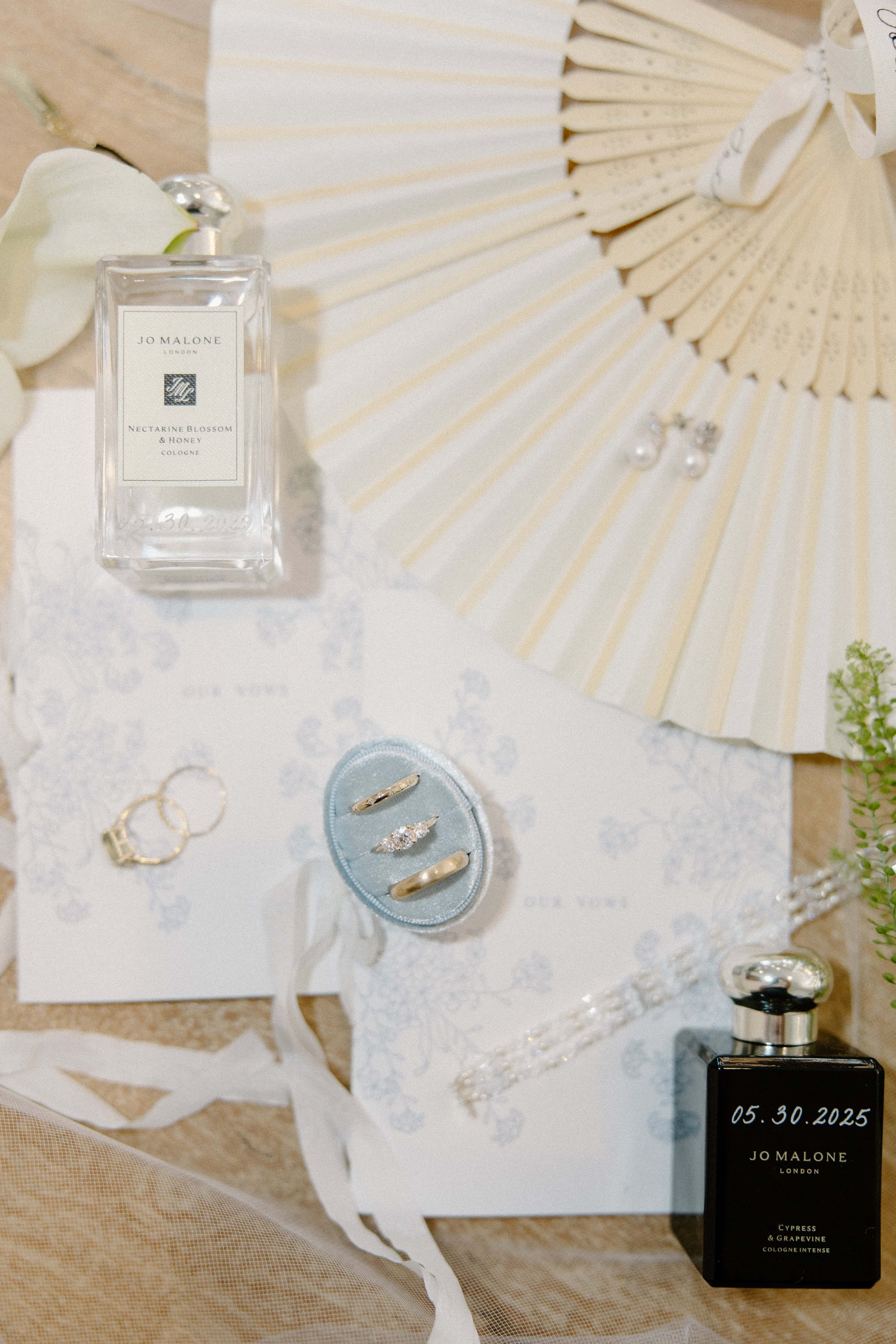 A flat lay photo of wedding accessories, including two bottles of perfume, a fan, and a ribbon with two rings tied to it.