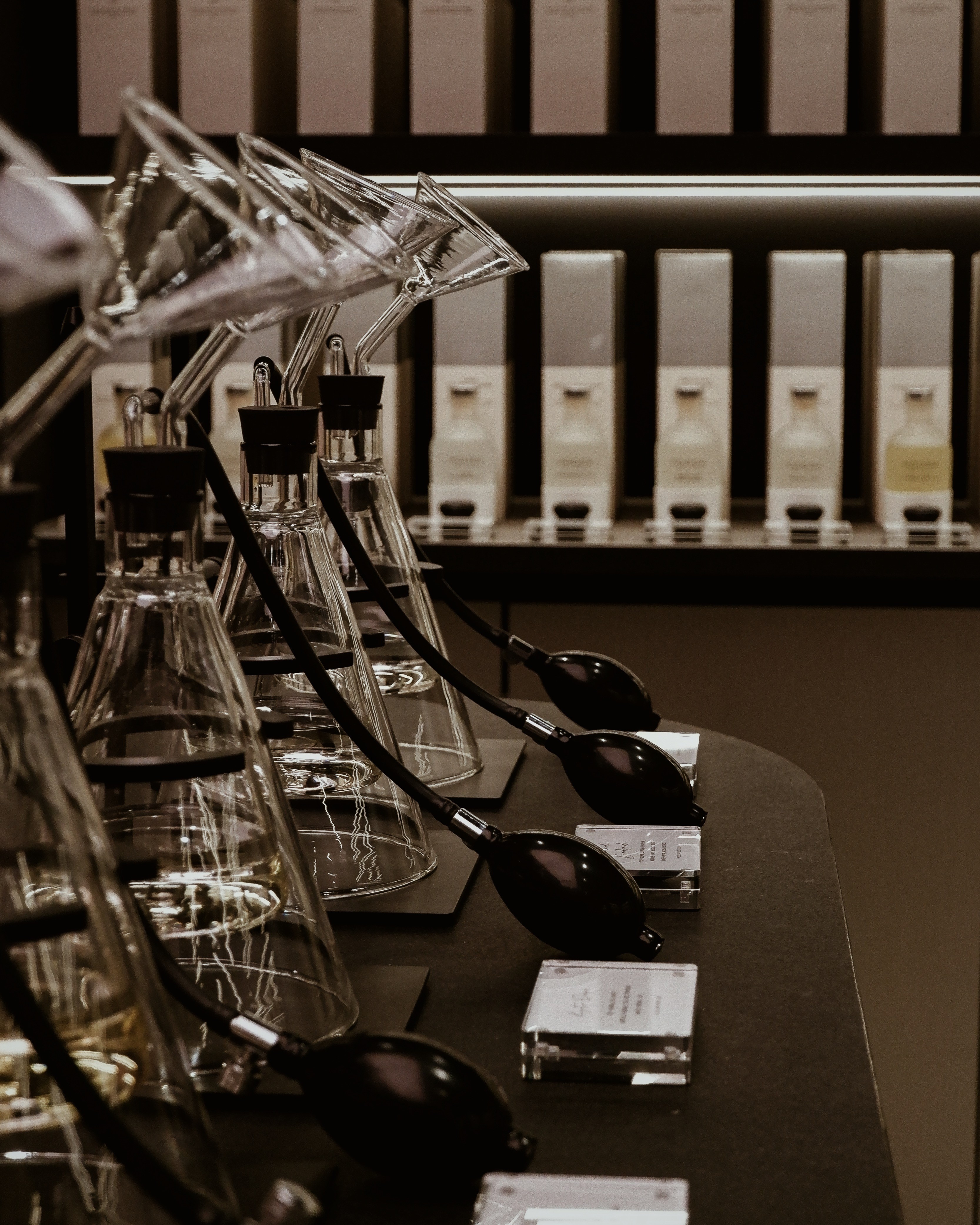 A close-up view of glass scent-testing equipment with black bulb syringes on a dark table, alongside perfume samples displayed in the background.