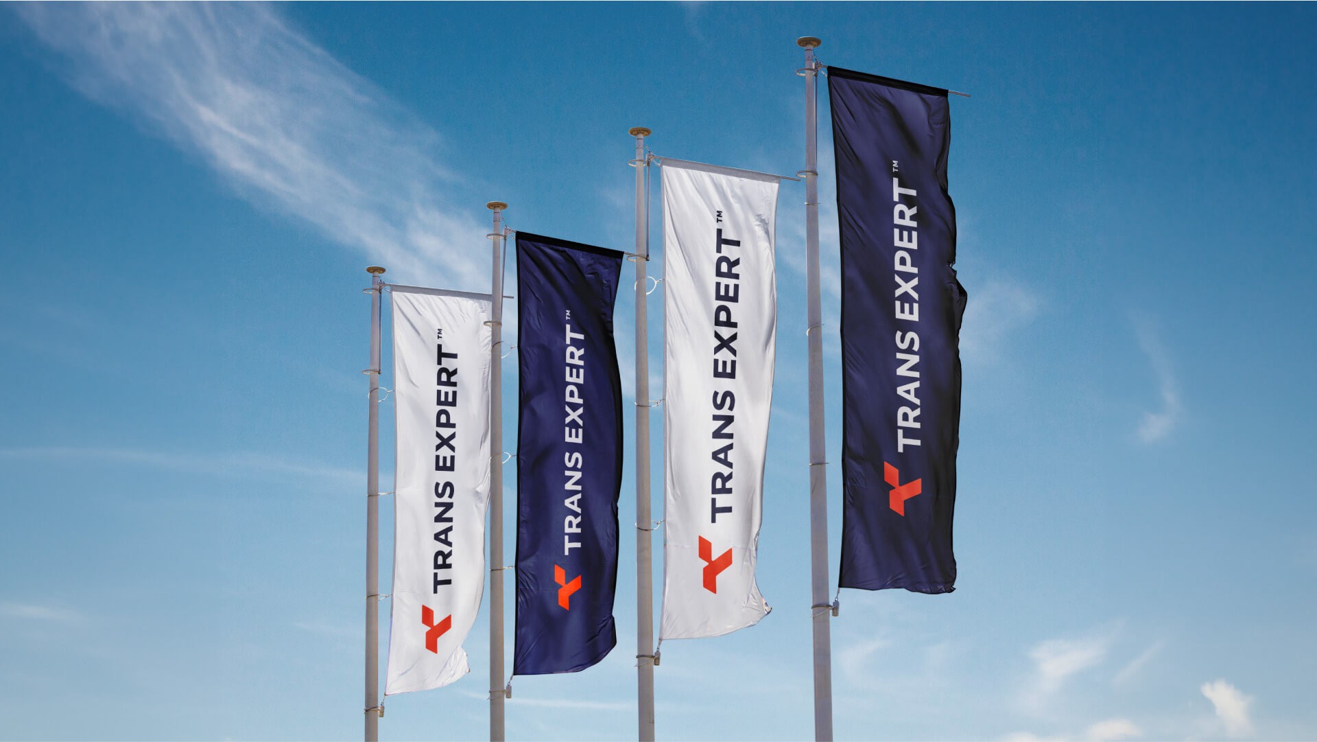 A square, illuminated exterior business sign for Trans Expert mounted on a building. The sign is dark navy blue with the prominent orange 'T' logo and 'TRANS EXPERT' text underneath.