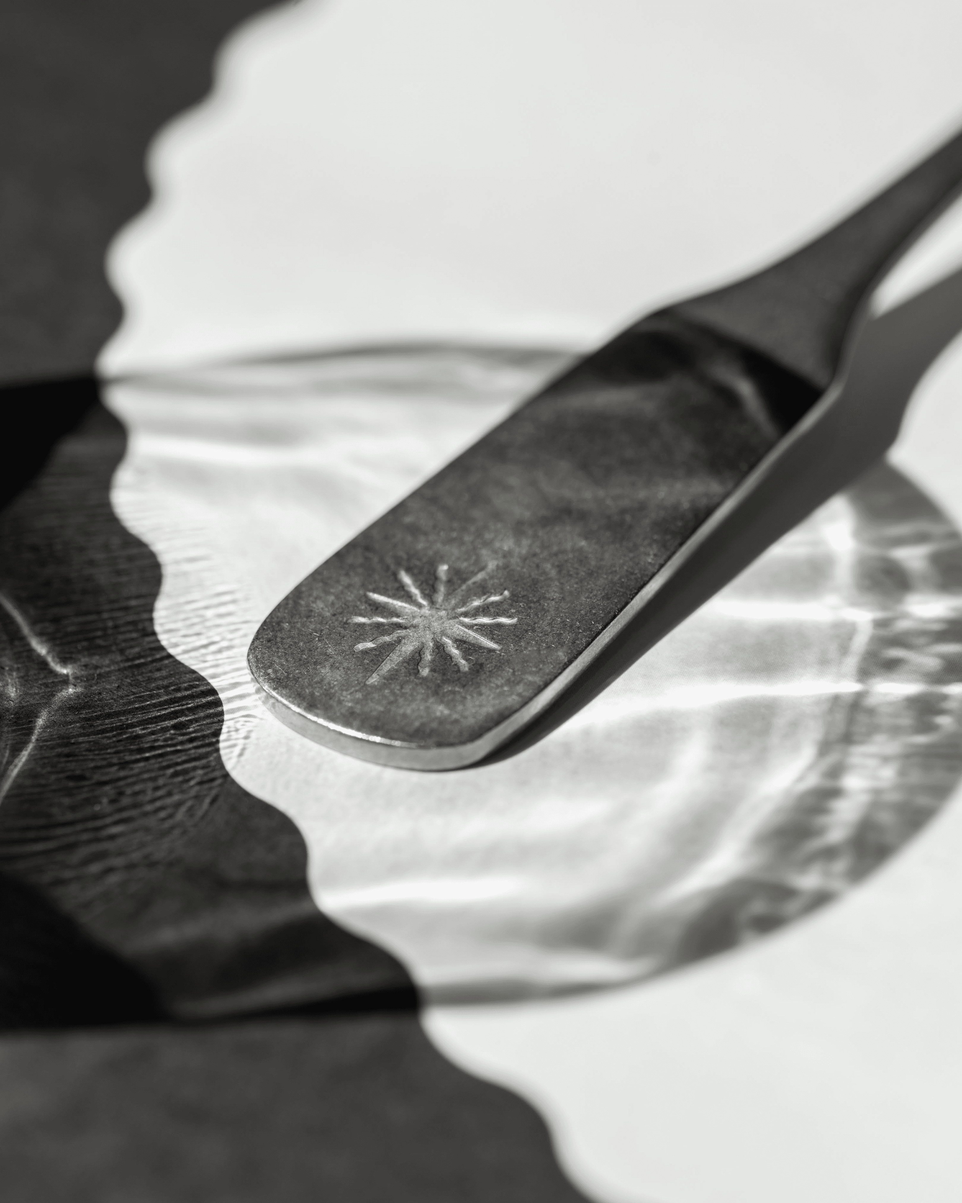 Close-up of matte black tweezers with star-shaped emblem on handle, gripping a metallic surface over reflective glass with soft shadows.
