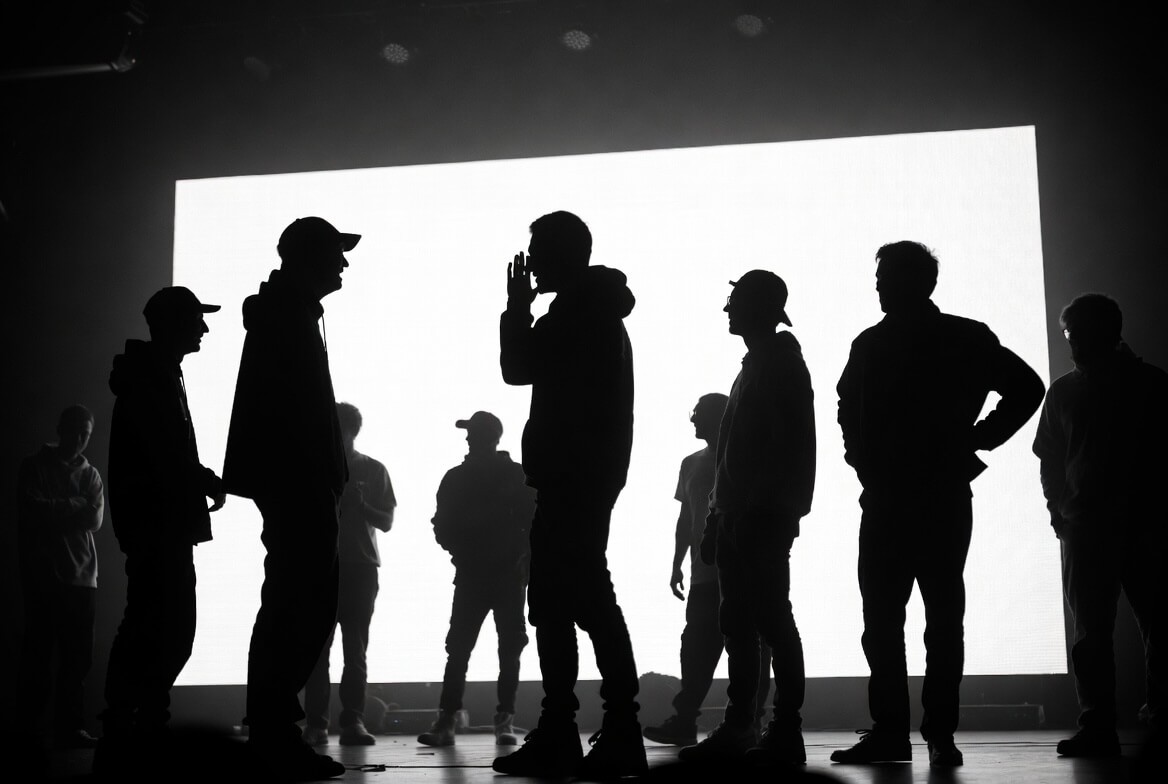 Silhouette of a creative team in a strategy meeting. One team member points at a large screen displaying project roadmap, flowcharts, and visual direction while others discuss and collaborate. High-contrast black and white backlit scene.