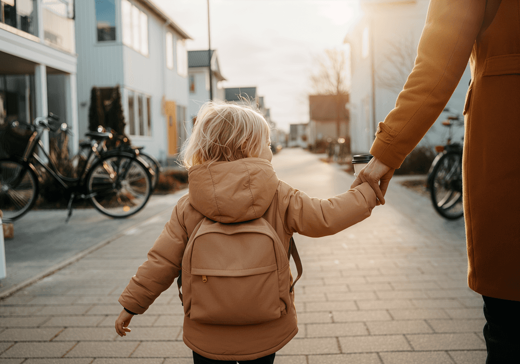 A child in a tan jacket holding an adult's hand, walking on a sidewalk. The child is wearing a backpack.